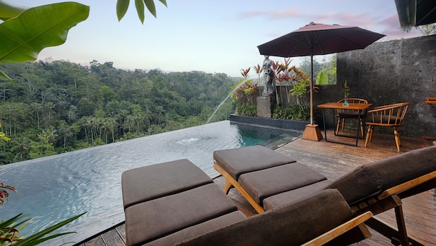 Una piscina infinita con sillones de descanso y una mesa, rodeada de exuberante vegetación y una estatua.