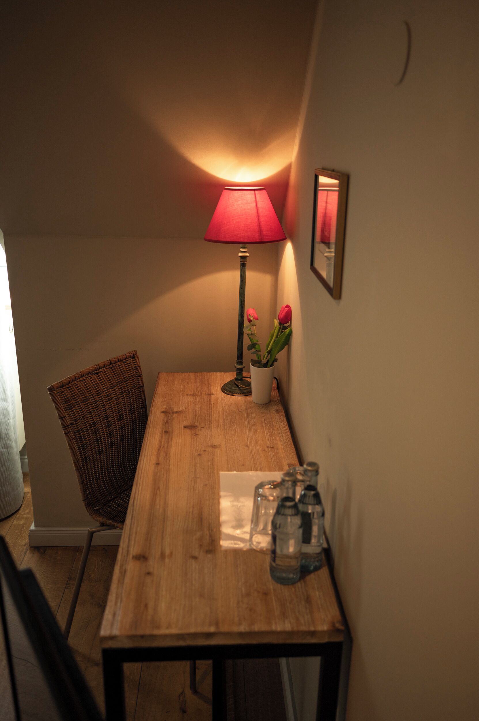 Un rincón de una habitación con una mesa de madera, una lámpara roja, una silla de mimbre, una planta en maceta con flores y un cuadro en la pared.