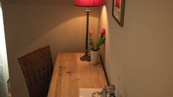 Un rincón de una habitación con una mesa de madera, una lámpara roja, una silla de mimbre, una planta en maceta con flores y un cuadro en la pared.