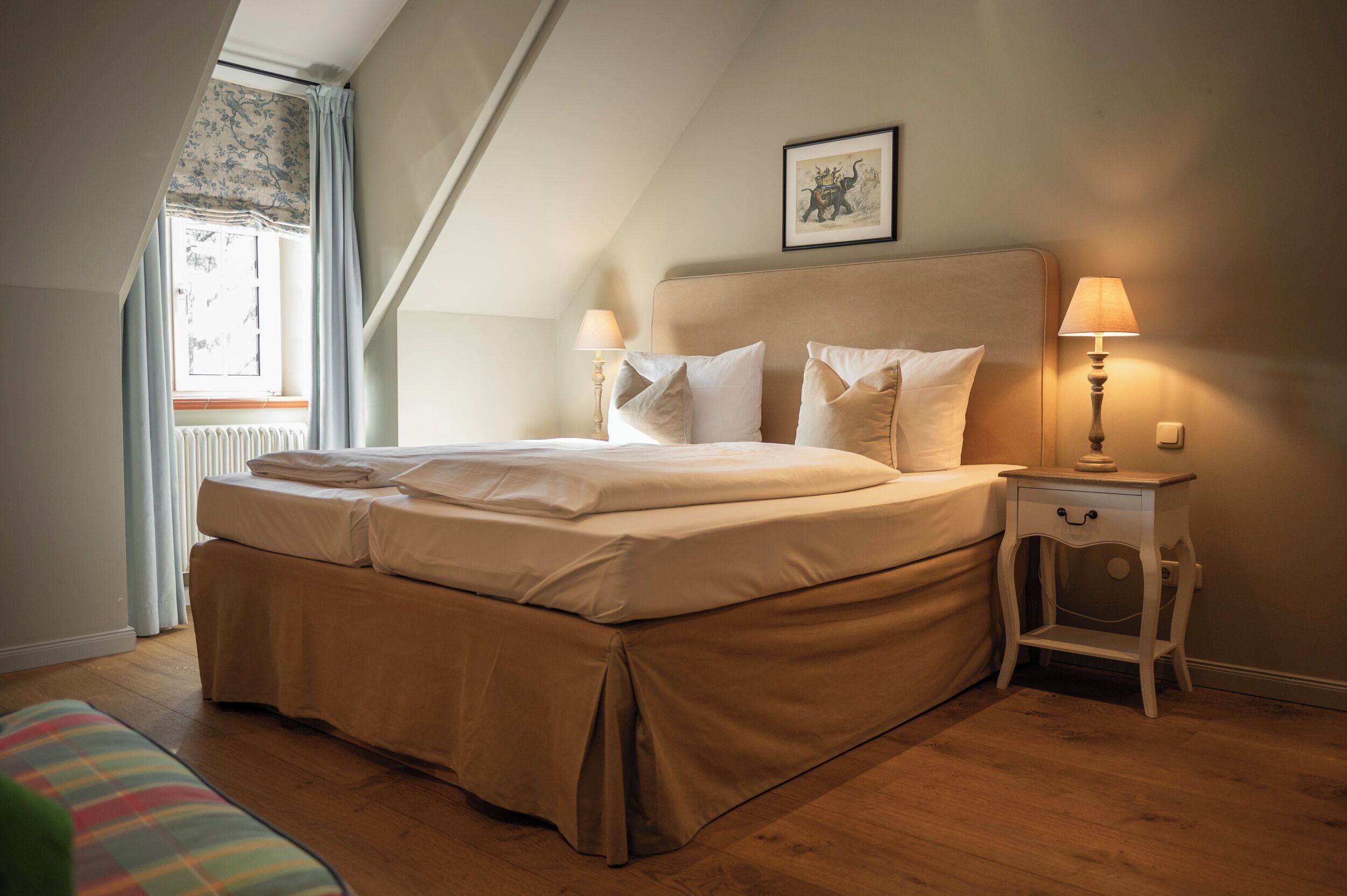 A bedroom with a large bed, two bedside tables with lamps, a window with curtains, and a framed picture on the wall.