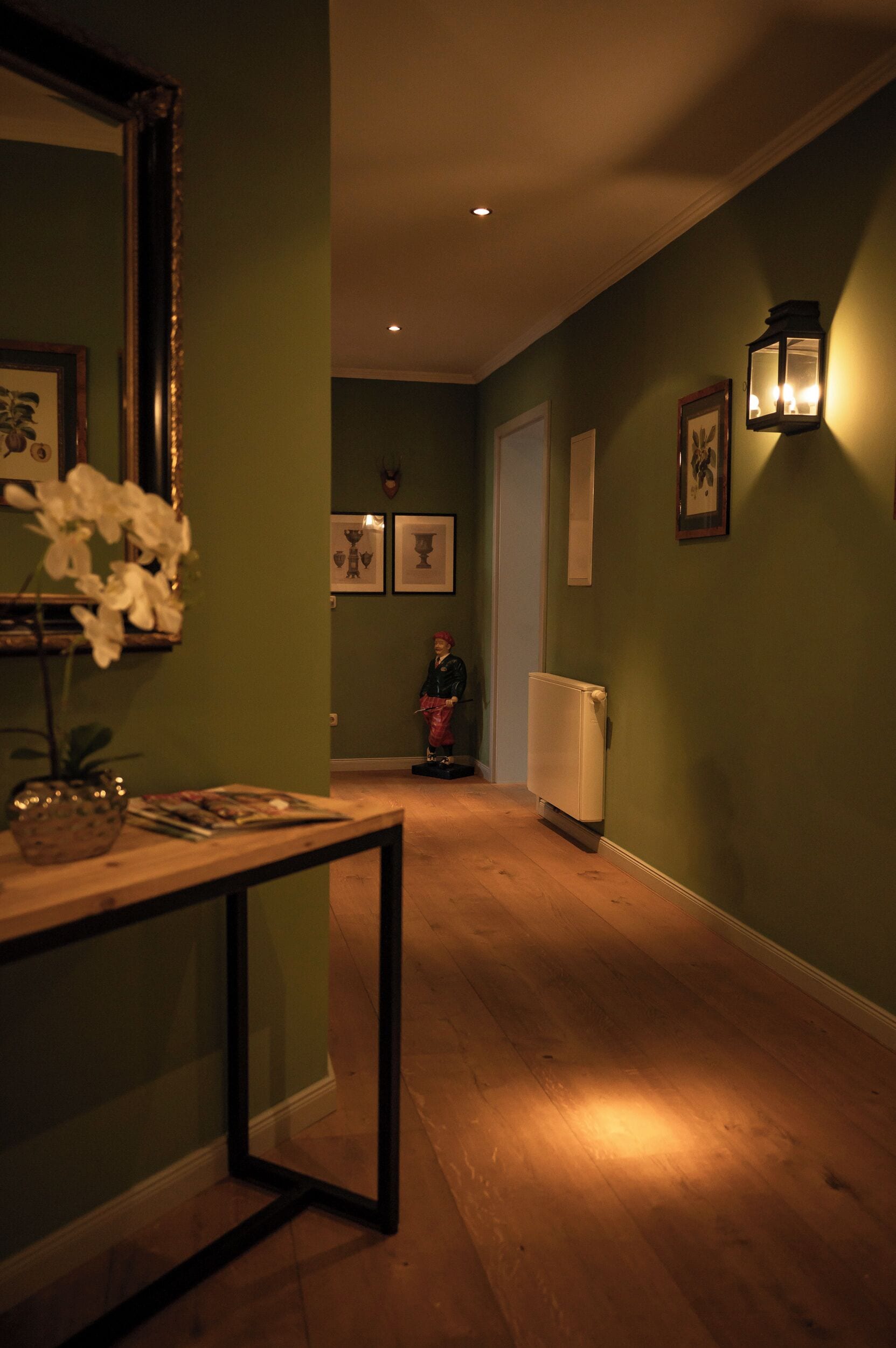 A hallway with a wooden floor, a table with a vase of flowers, a mirror, and framed pictures on the walls.