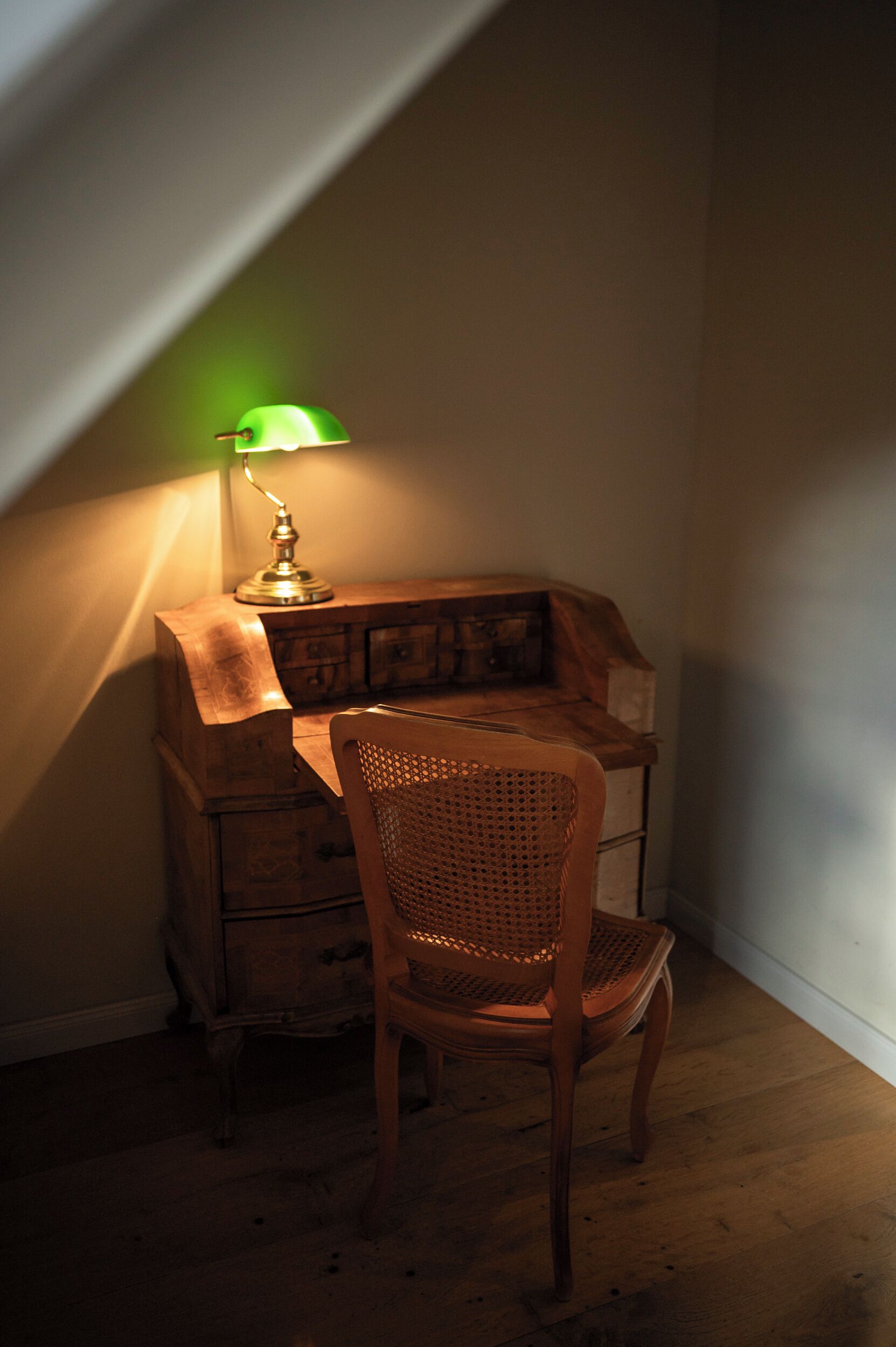 A wooden desk with a lamp, a wicker chair, and a wooden chest of drawers in a corner of a room.