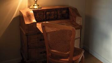 A wooden desk with a lamp, a wicker chair, and a wooden chest of drawers in a corner of a room.