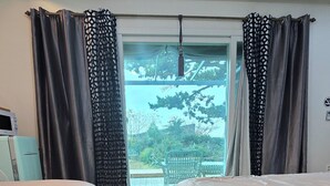 A hotel room with two beds, a large window with curtains, a ceiling fan, and a view of greenery outside. - Wind Jeju Cafe & Pension (Seogwipo)