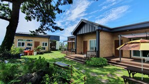 A modern two-story house with a patio, a barbecue area, and a deck with outdoor furniture. - Wind Jeju Cafe & Pension (Seogwipo)