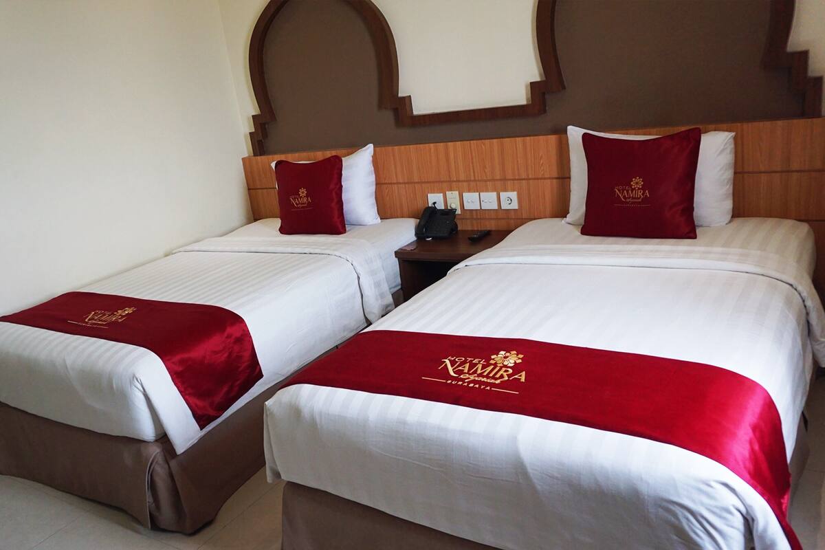 Habitación de hotel con dos camas, cada una con sábanas blancas y una colcha roja que muestra el nombre del hotel. Se observa un cabecero de madera, una mesita de noche con teléfono y control remoto, y un interruptor de luz en la pared.