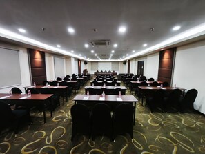 Uma sala de conferência com fileiras de mesas e cadeiras, equipada com garrafas de água, e uma tela de projeção na frente.