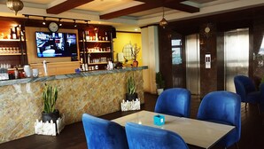 Um saguão de hotel com uma mesa de recepção, cadeiras azuis, um relógio e uma televisão exibindo uma cena de bar.
