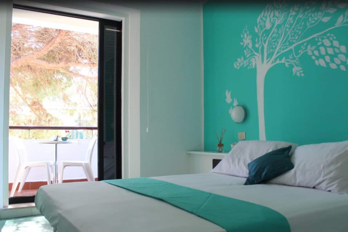 Een slaapkamer met een bed, nachtkastje, muurschildering en een balkon met stoelen.