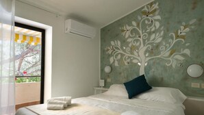 A hotel room with a bed, a window with a view of greenery, a wall with a floral mural, and an air conditioning unit.