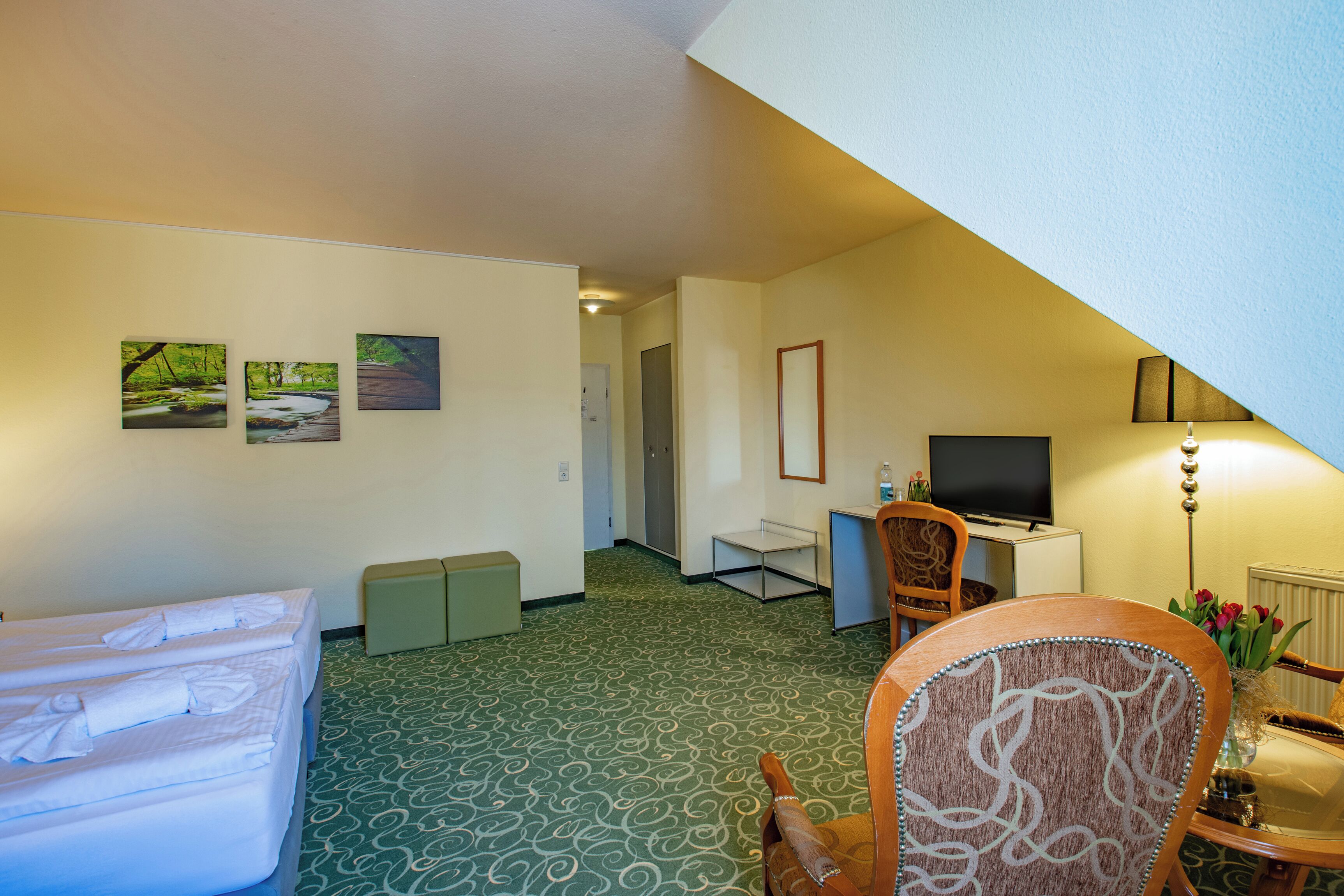 A hotel room with a bed, a desk, a chair, a TV, and a small table with a vase of flowers.