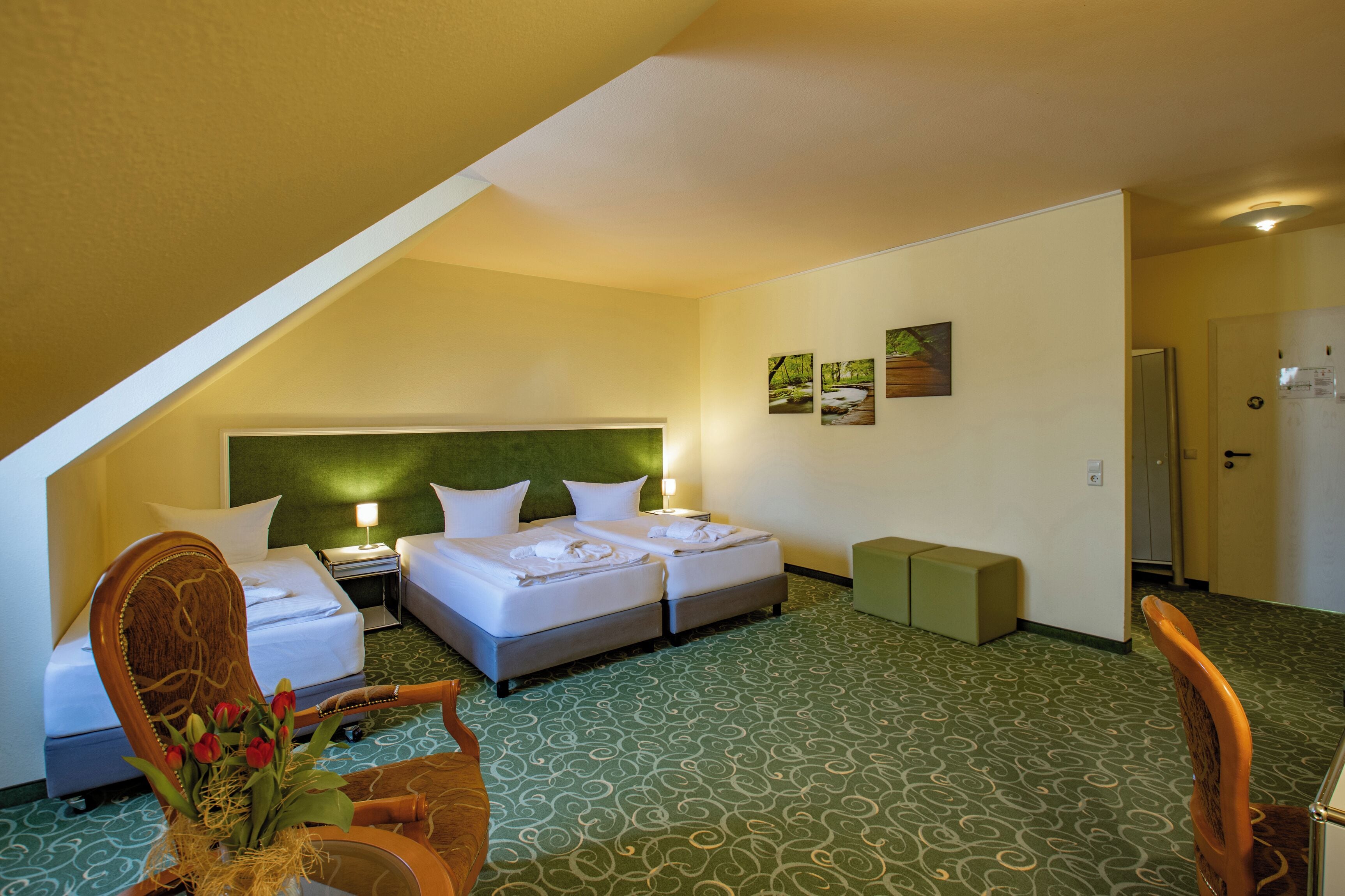 Habitación de hotel con dos camas, alfombra verde estampada, una silla de madera y una mesita con un jarrón de flores.