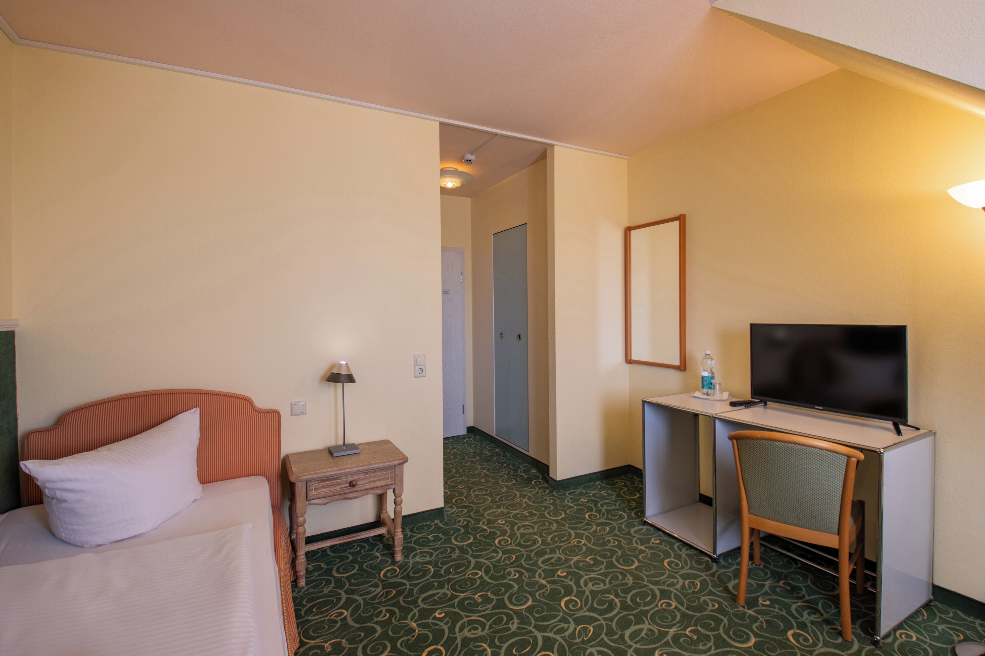 Et hotelværelse med en seng, et skrivebord med fjernsyn, en stol og et lille bord med lampe.