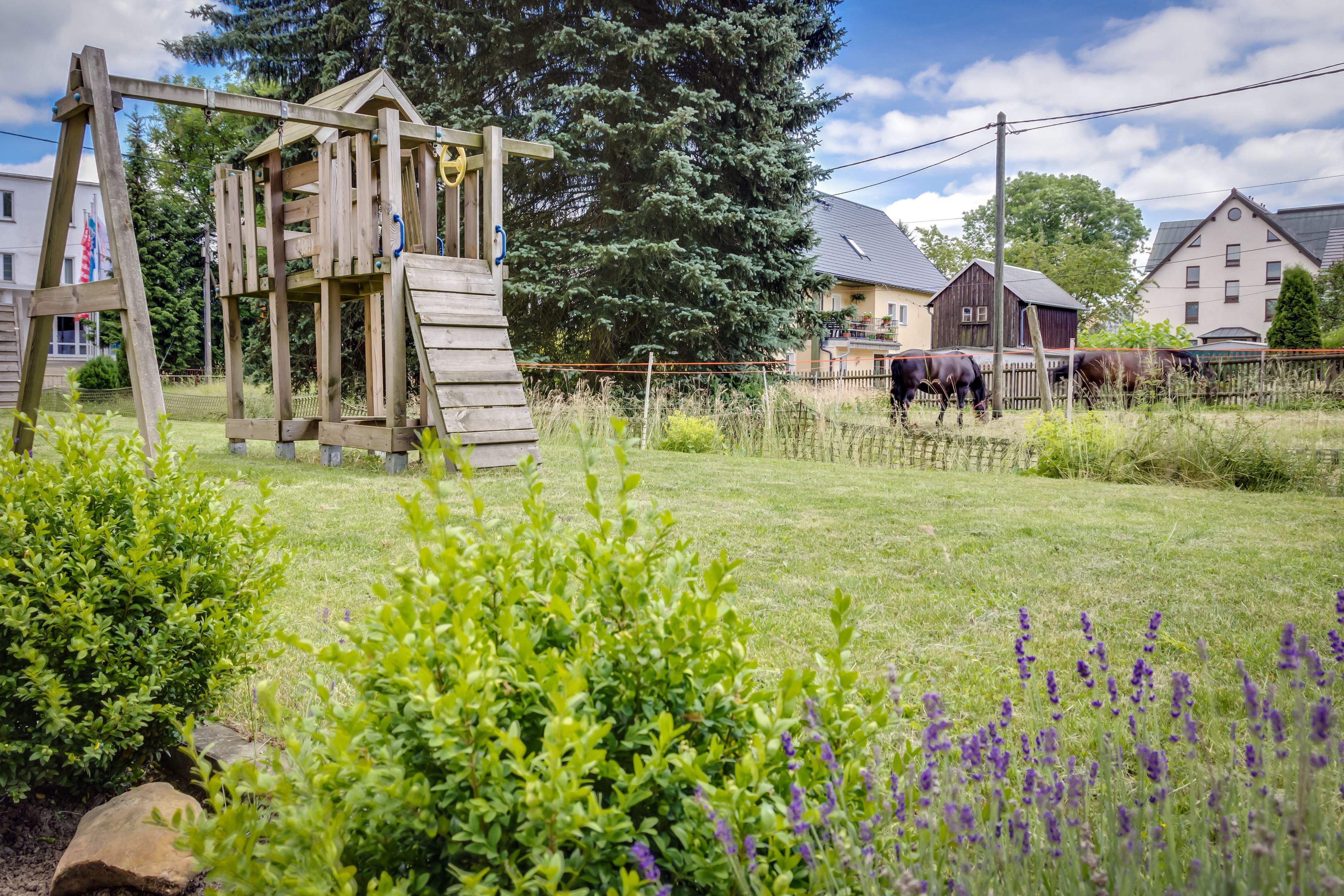 Une structure de jeu en bois, installée dans une cour verdoyante, avec des maisons en arrière-plan.