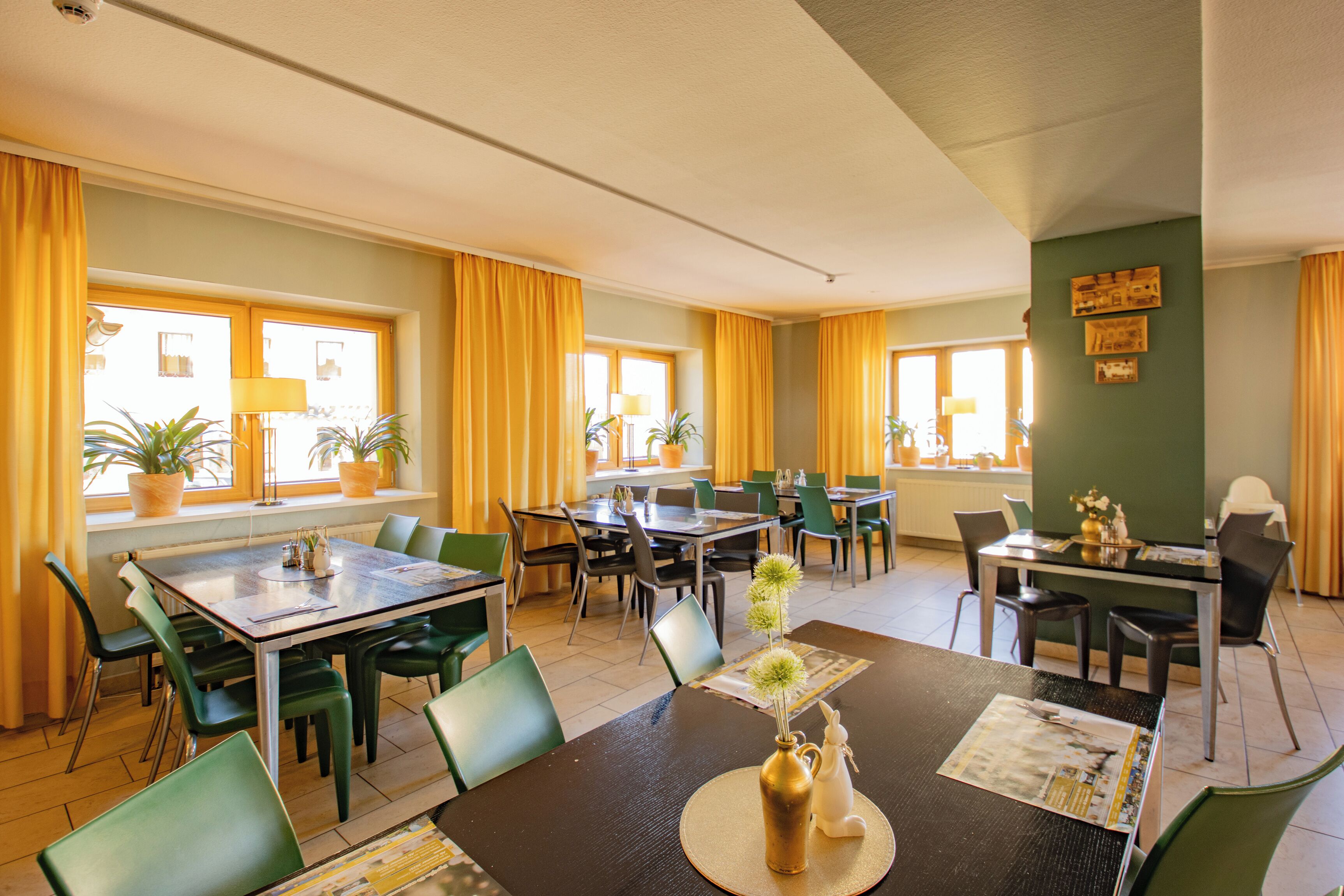 A hotel dining area with multiple tables and chairs, potted plants, and a view of the street through large windows.
