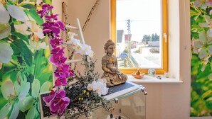 Una estatua de una figura sentada sobre una estantería, rodeada de orquídeas, con una ventana al fondo.