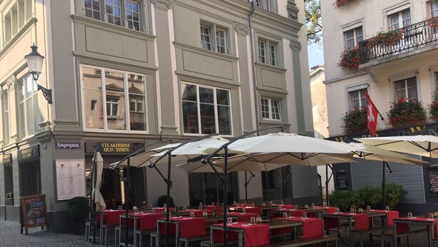 An outdoor café with tables and umbrellas, a sign for 'Steakhouse Old Town', and a building with a flag and flower boxes. - Hotel Old Town (Zürich)