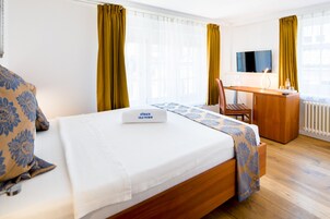 Quarto de hotel com cama, escrivaninha, cadeira, televisão e janela com cortinas.