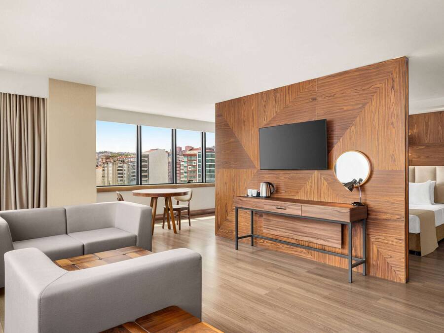 A modern hotel room with a large window, a grey sofa, a wooden accent wall, a flat-screen TV, and a bed.