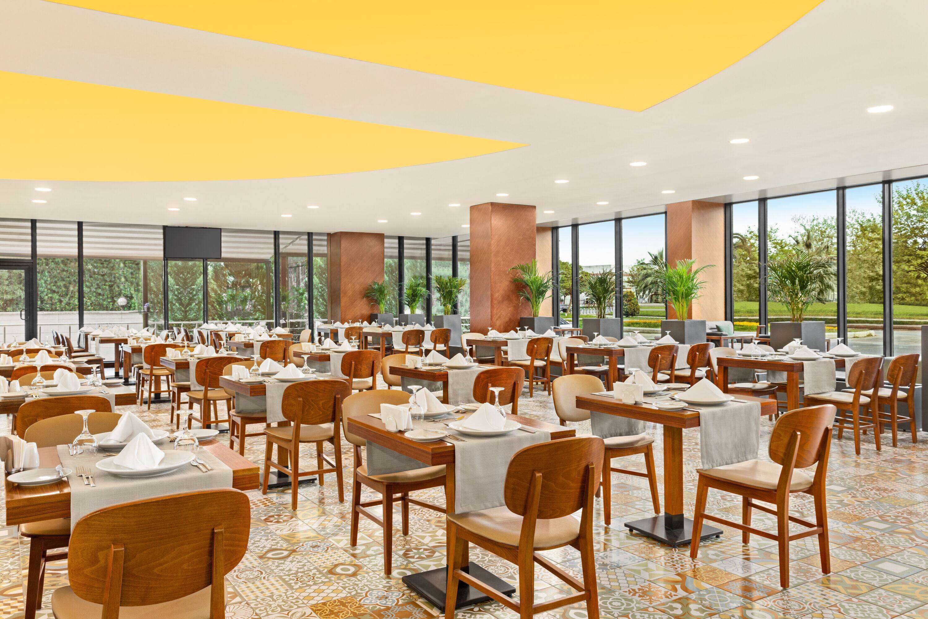A spacious dining area with multiple tables and chairs, large windows offering views of greenery, and a modern ceiling design.
