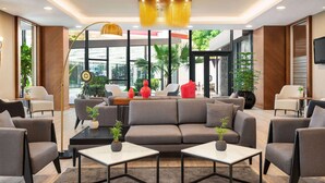 Un vestíbulo moderno con un sofá gris, sillones y una mesa de centro. Hay plantas en macetas y un ventanal con vistas a palmeras.