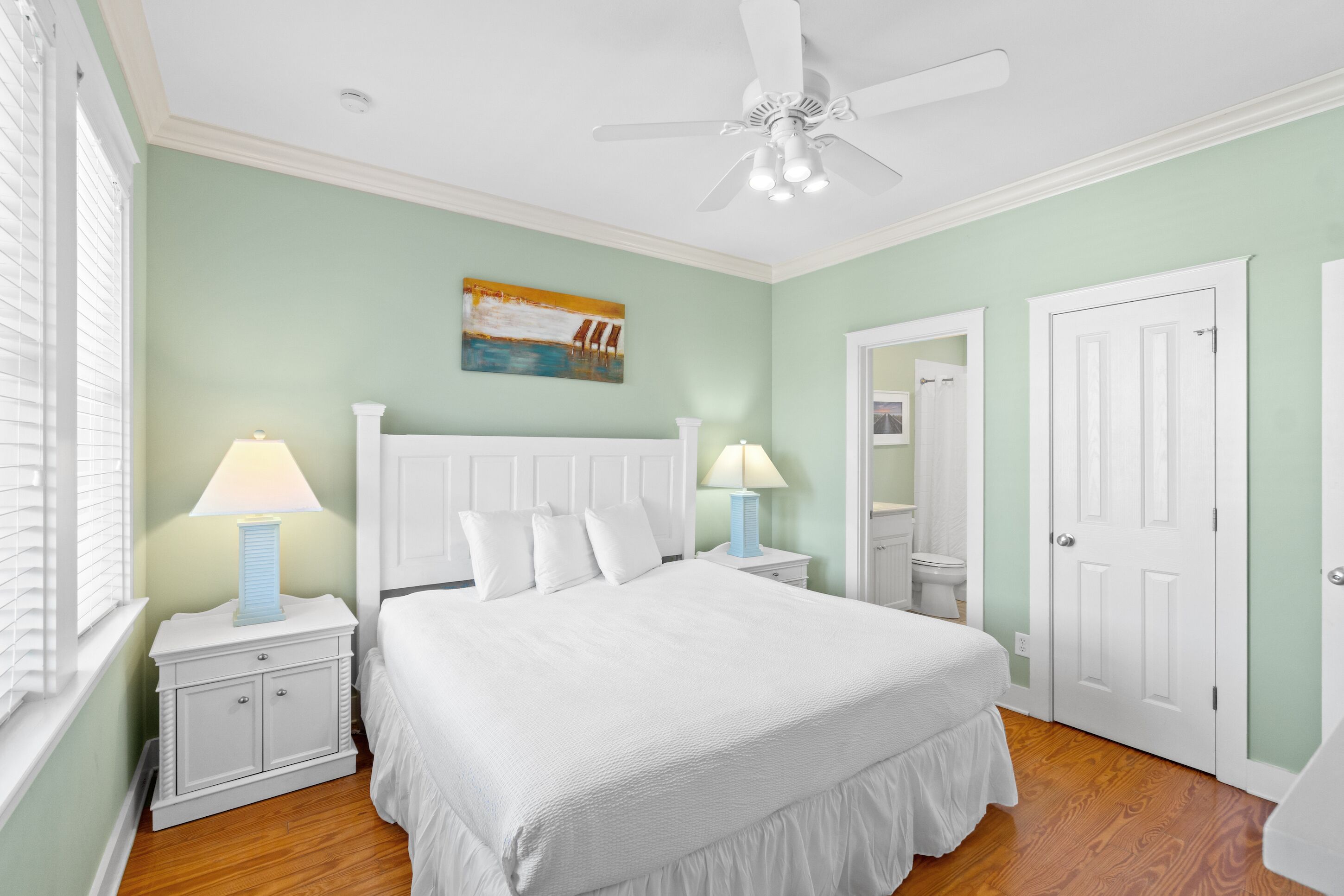 A bedroom with a white bed, two bedside tables with lamps, a ceiling fan, and a painting on the wall.