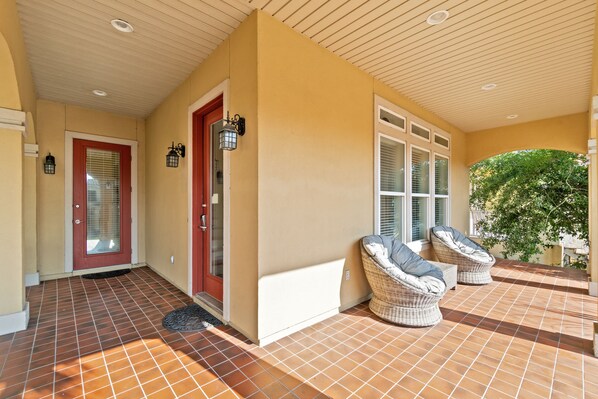 A covered porch with two wicker chairs, a red door, and a tiled floor.
