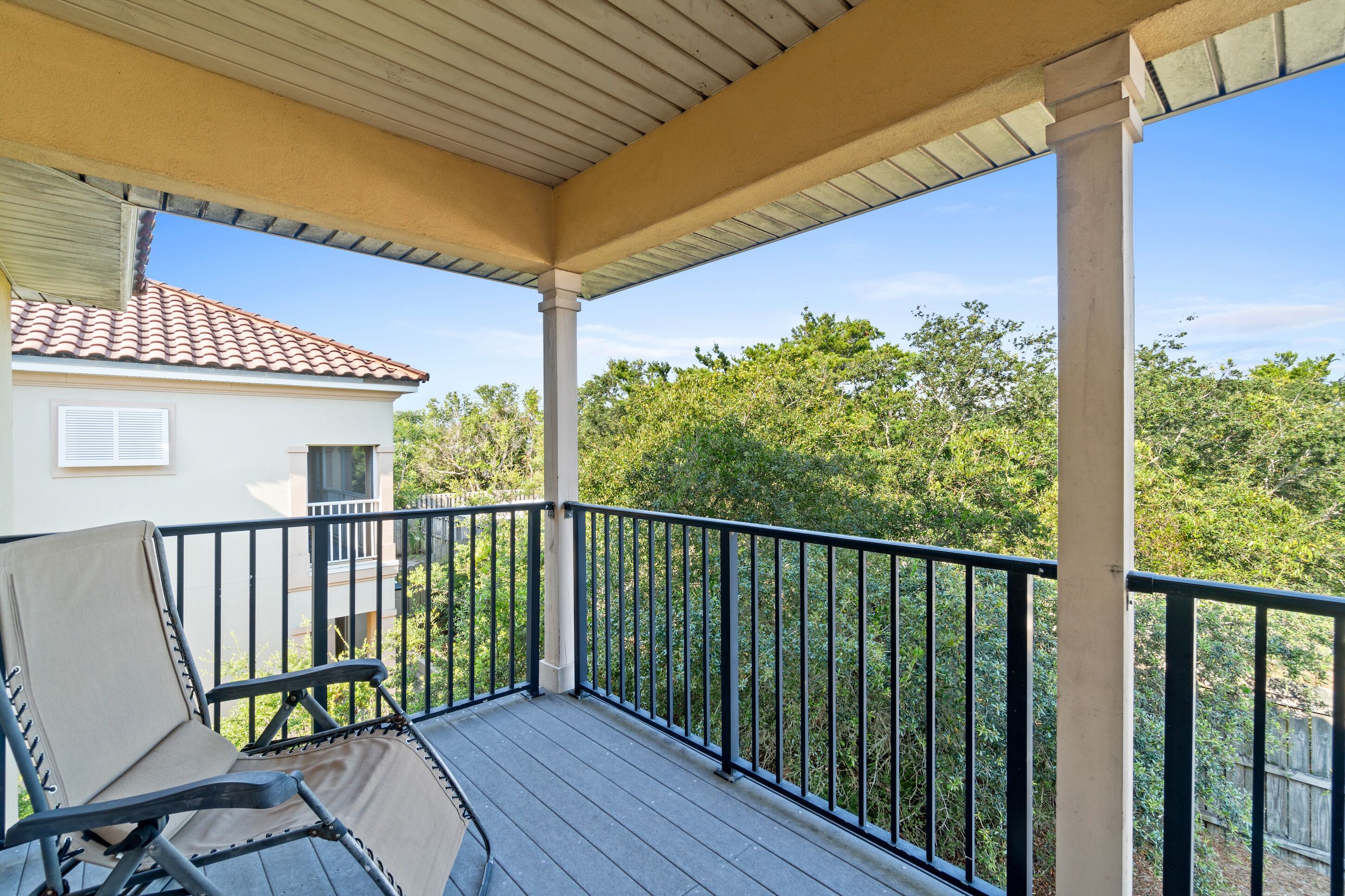 A covered balcony with a chair, a railing, and a view of a house and trees.