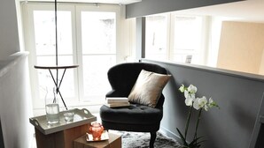 A modern living room with a black armchair, a wooden side table, a glass coffee table, a potted orchid, and a large window with natural light. - Lille A Part - Gites Urbains (Lille)