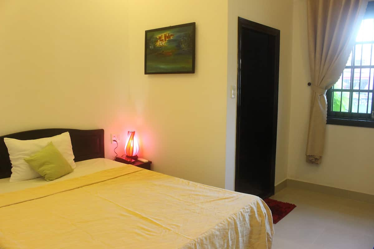 A neatly made bed with a white bedspread and pillows, a bedside table with a lamp, a painting on the wall, and a window with curtains.