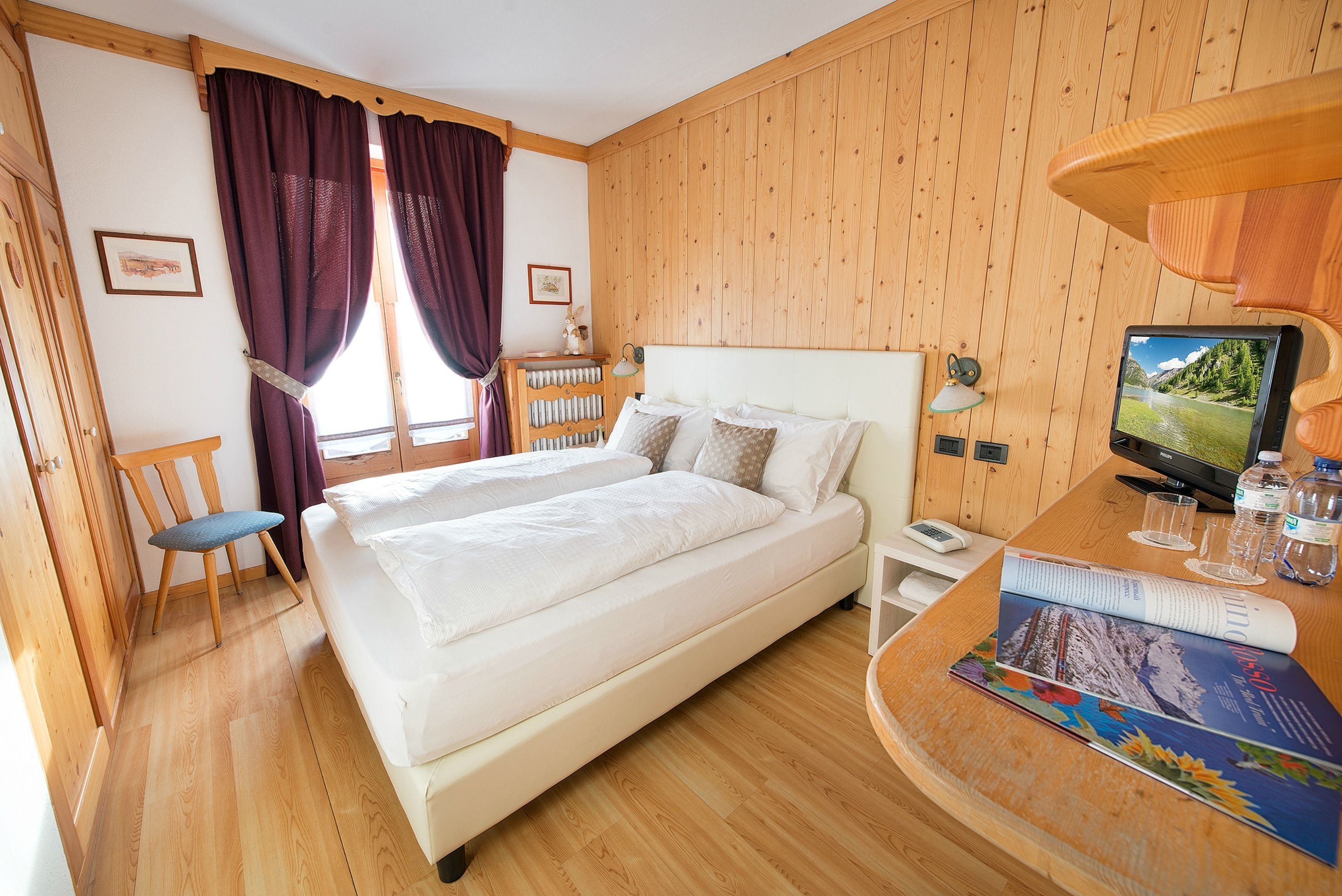 A hotel room with a wooden floor, a bed with white bedding, a desk with a computer and a magazine, and a window with curtains.