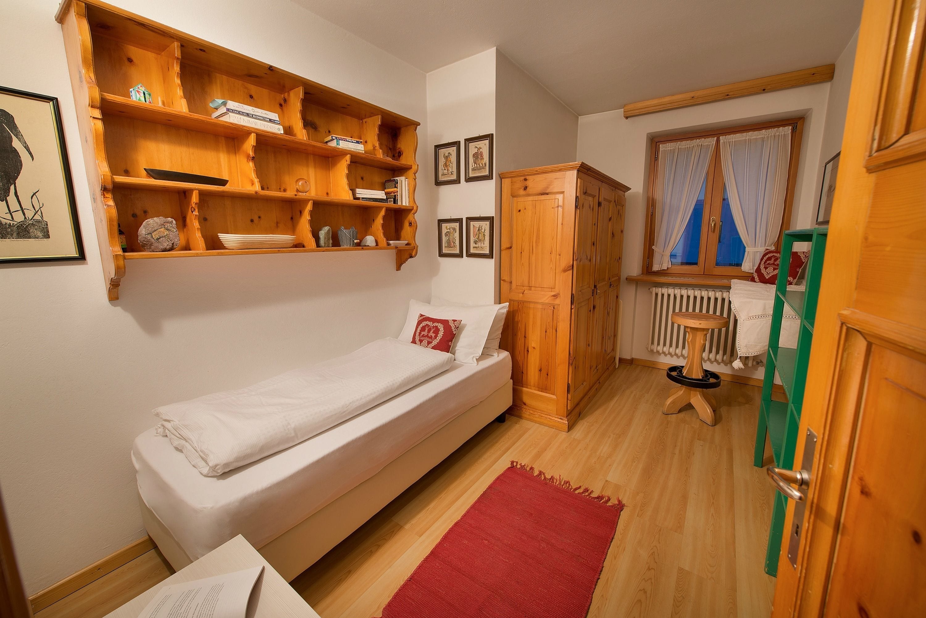 A small, neatly organized bedroom with a single bed, wooden shelves above, a radiator, and a window with curtains.
