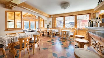 A dining area with wooden tables and chairs, large windows, and a tiled floor.