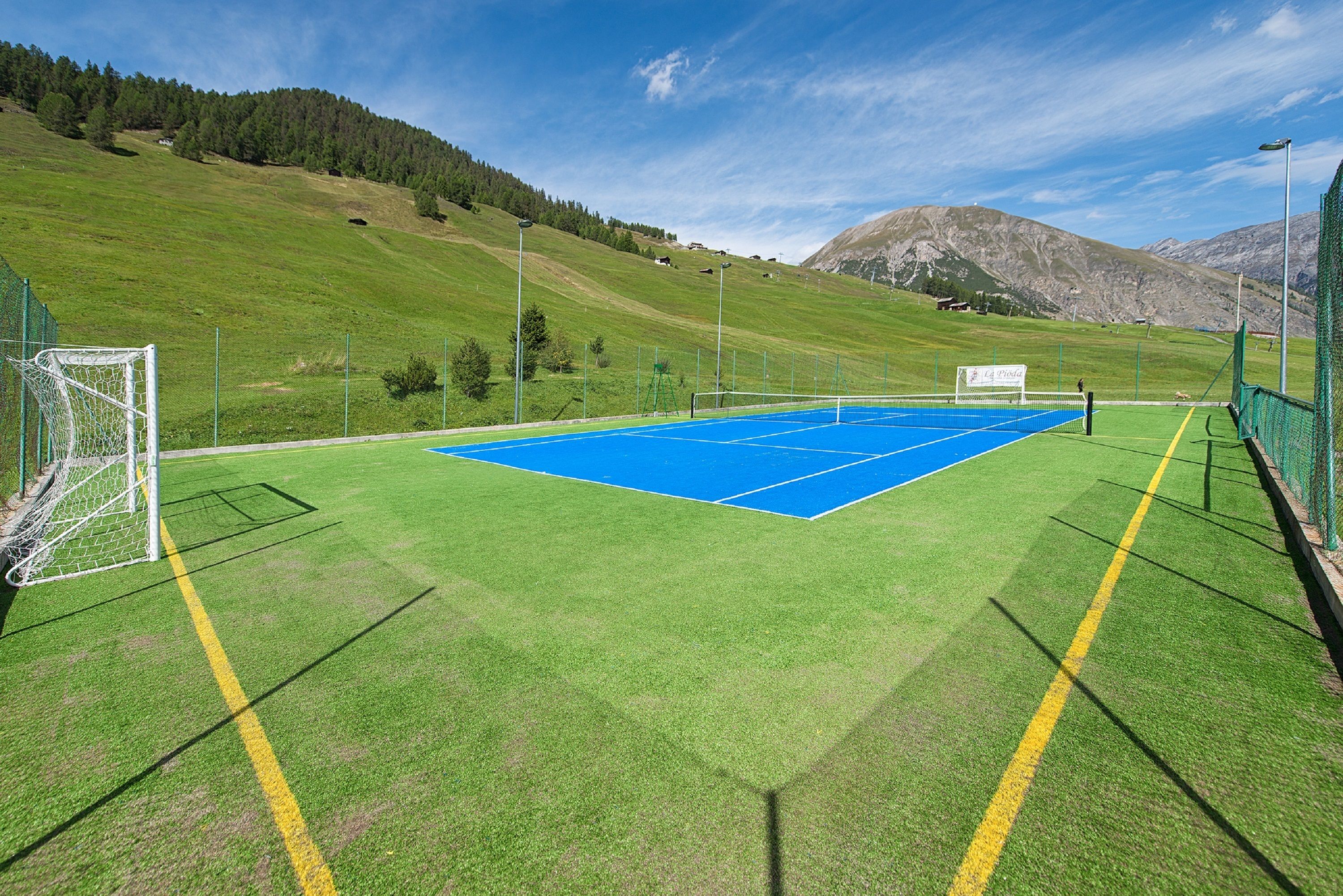 An outdoor tennis court with a goalpost, surrounded by green grass and mountains in the background.