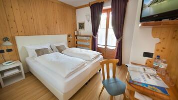 Quarto de hotel com duas camas, uma cabeceira de madeira, uma escrivaninha com cadeira, uma televisão e uma janela com cortinas.