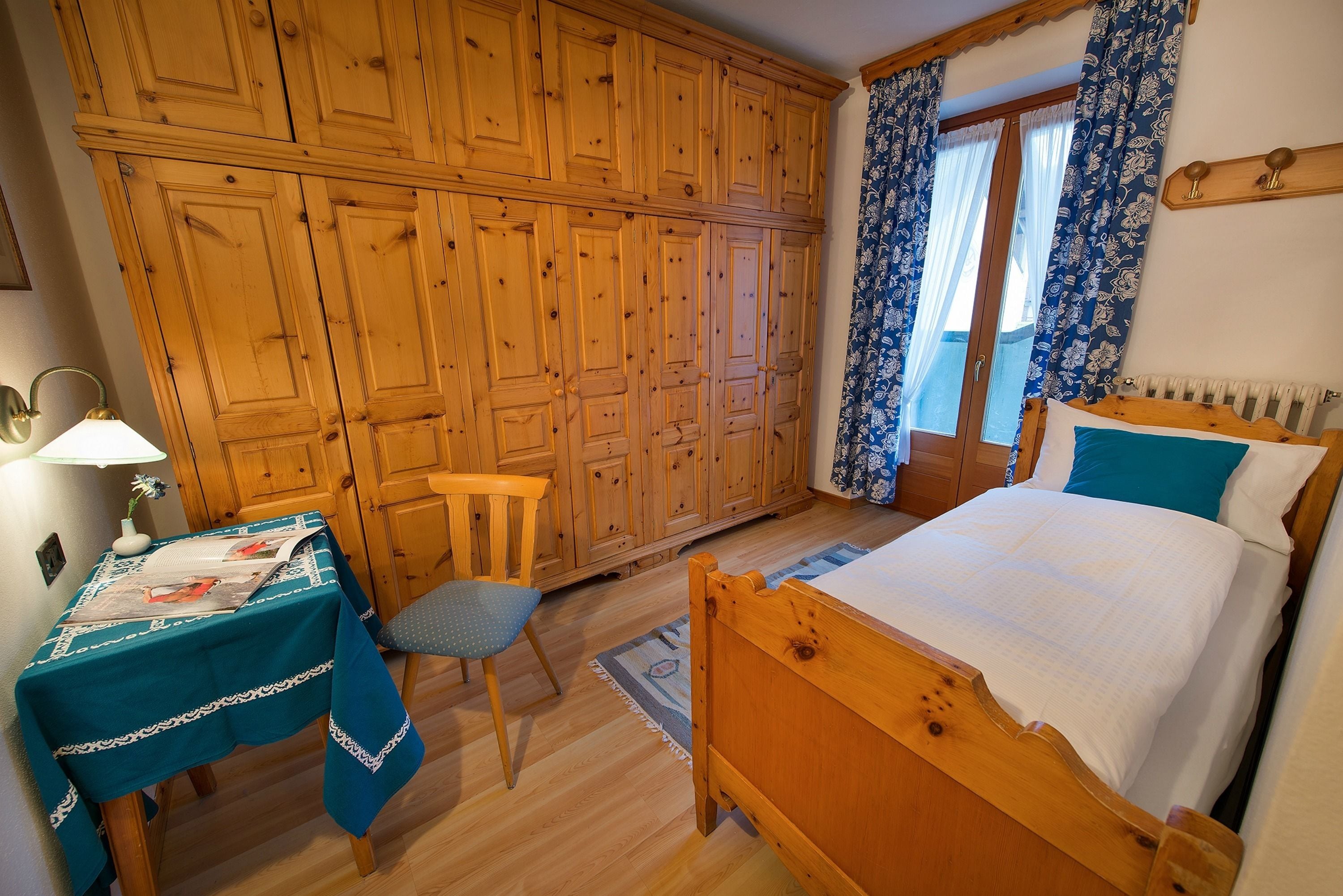 A bedroom with a wooden wardrobe, a bed with white bedding, a small table with a blue tablecloth, and a chair.
