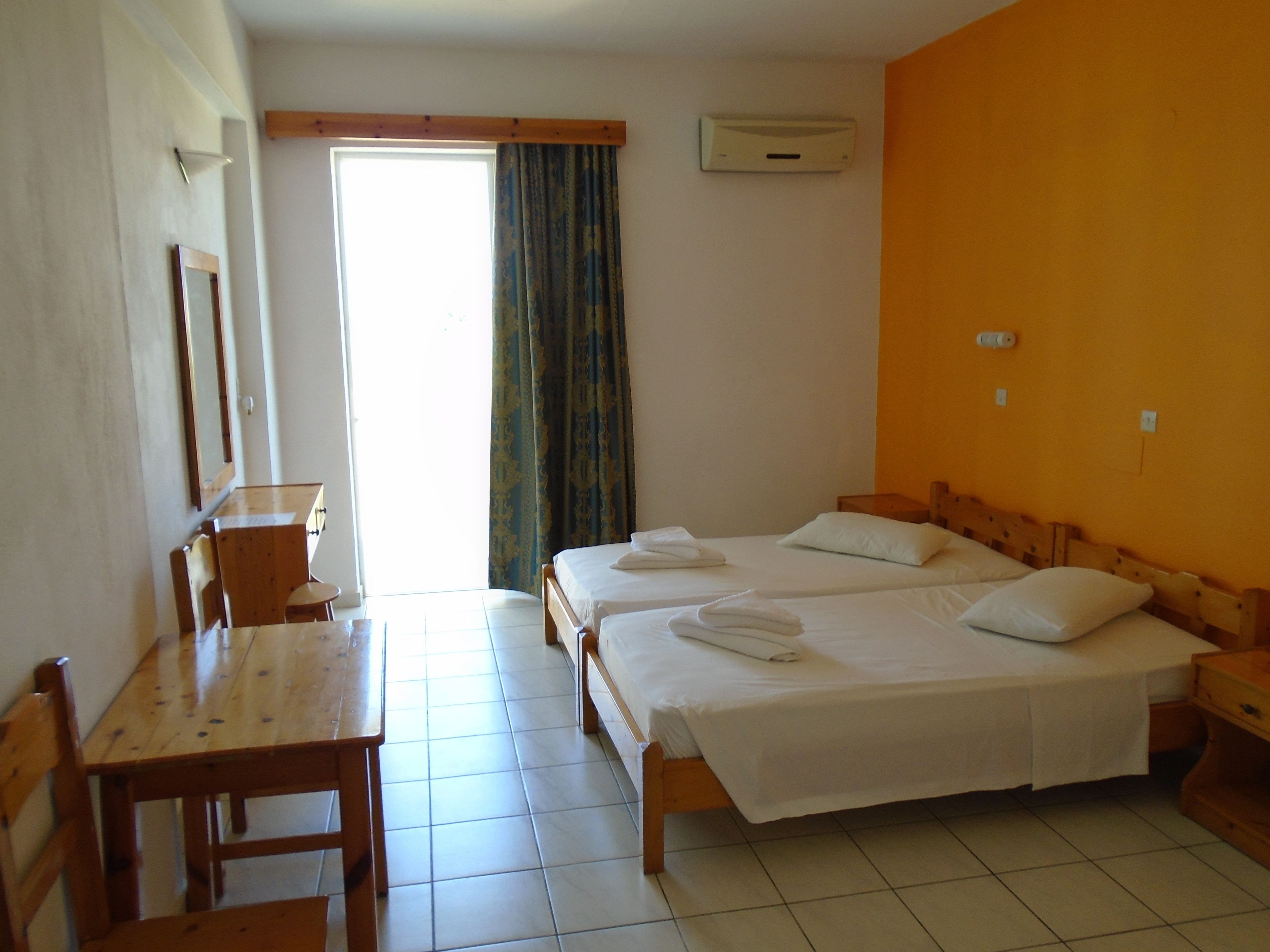 A hotel room with two beds, a wooden desk, a chair, a window with curtains, and an air conditioning unit.