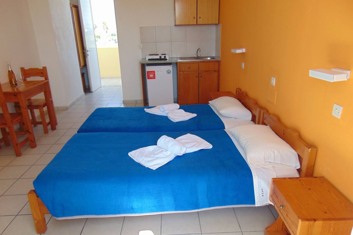 A hotel room with two beds, a wooden nightstand, a small kitchenette, and a dining area with a table and chairs.