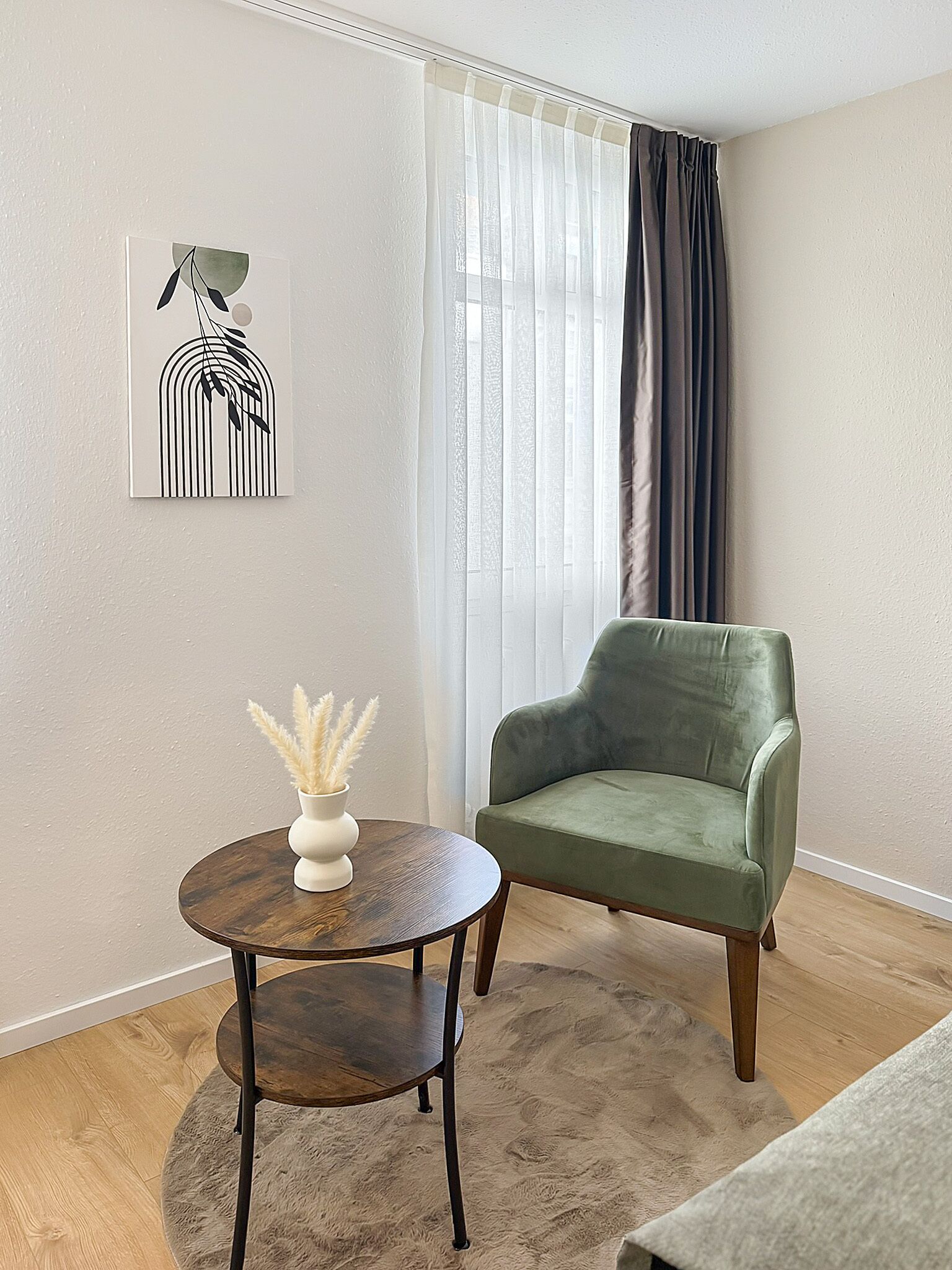 Una habitación con un sillón verde, una mesa redonda de madera con un jarrón de plumas de misel, y una obra de arte abstracto colgada en la pared.