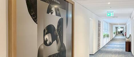 A hallway with a large framed artwork on the left, a carpeted floor, and a long corridor with windows and plants.