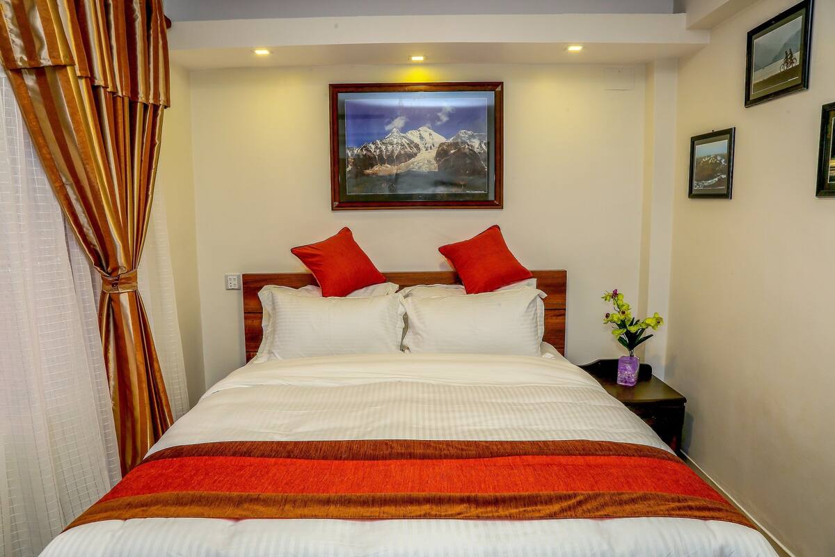 A neatly made bed with white and red bedding, a framed picture of mountains on the wall, and a small vase with flowers on the nightstand.