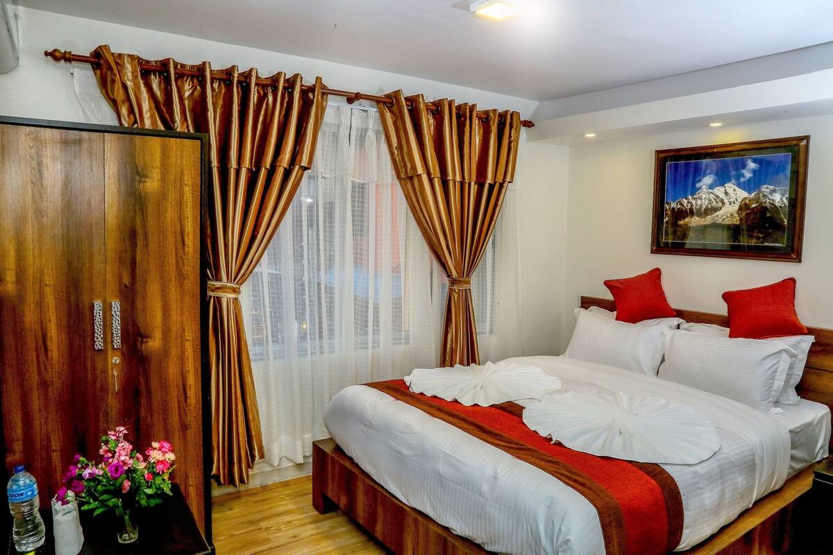 A hotel room with a wooden wardrobe, a bed with white bedding and red accents, a painting on the wall, and a window with sheer curtains.