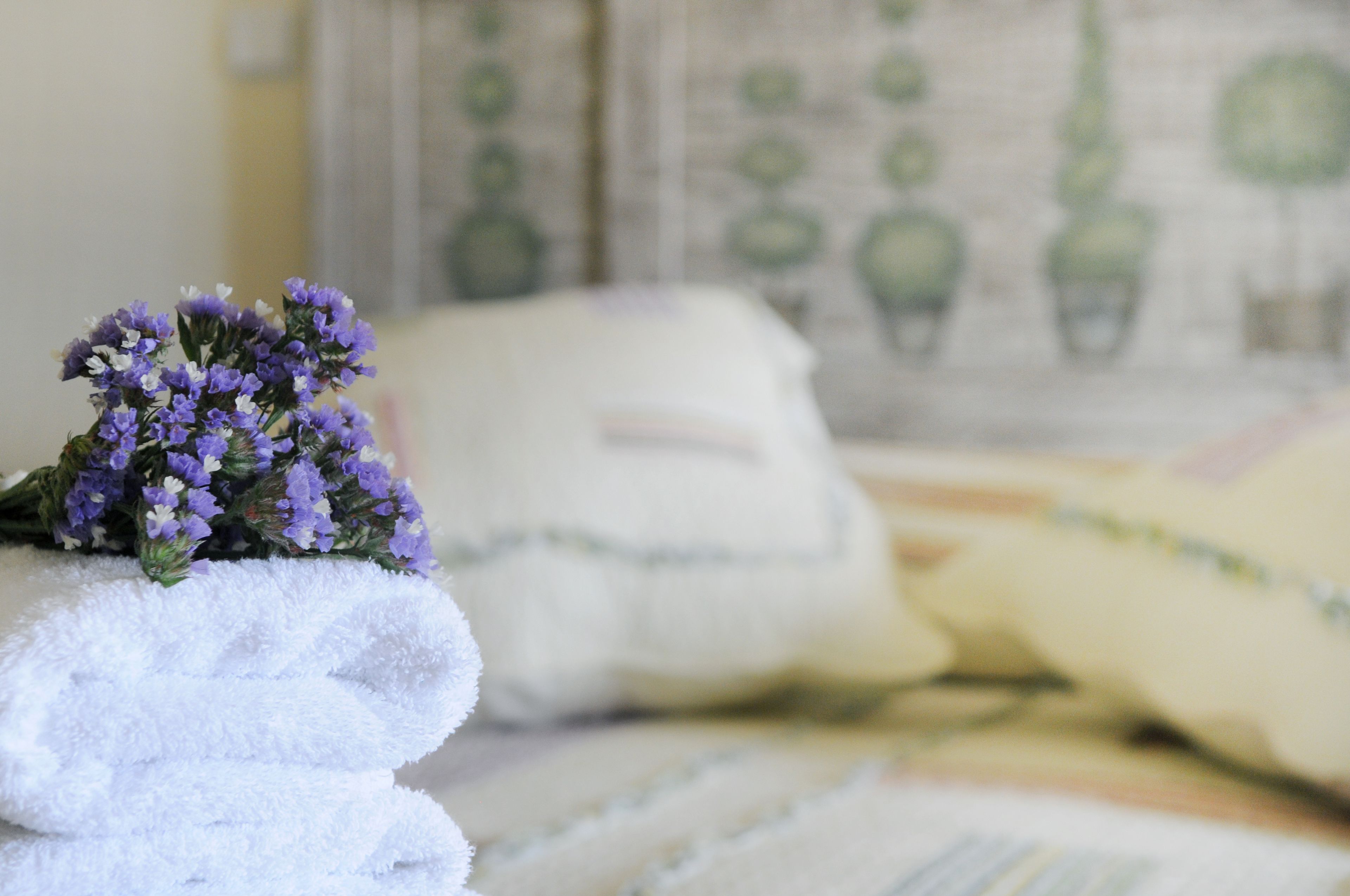 Una toalla blanca doblada con cuidado y un ramo de flores moradas sobre una cama con almohada y manta estampadas.