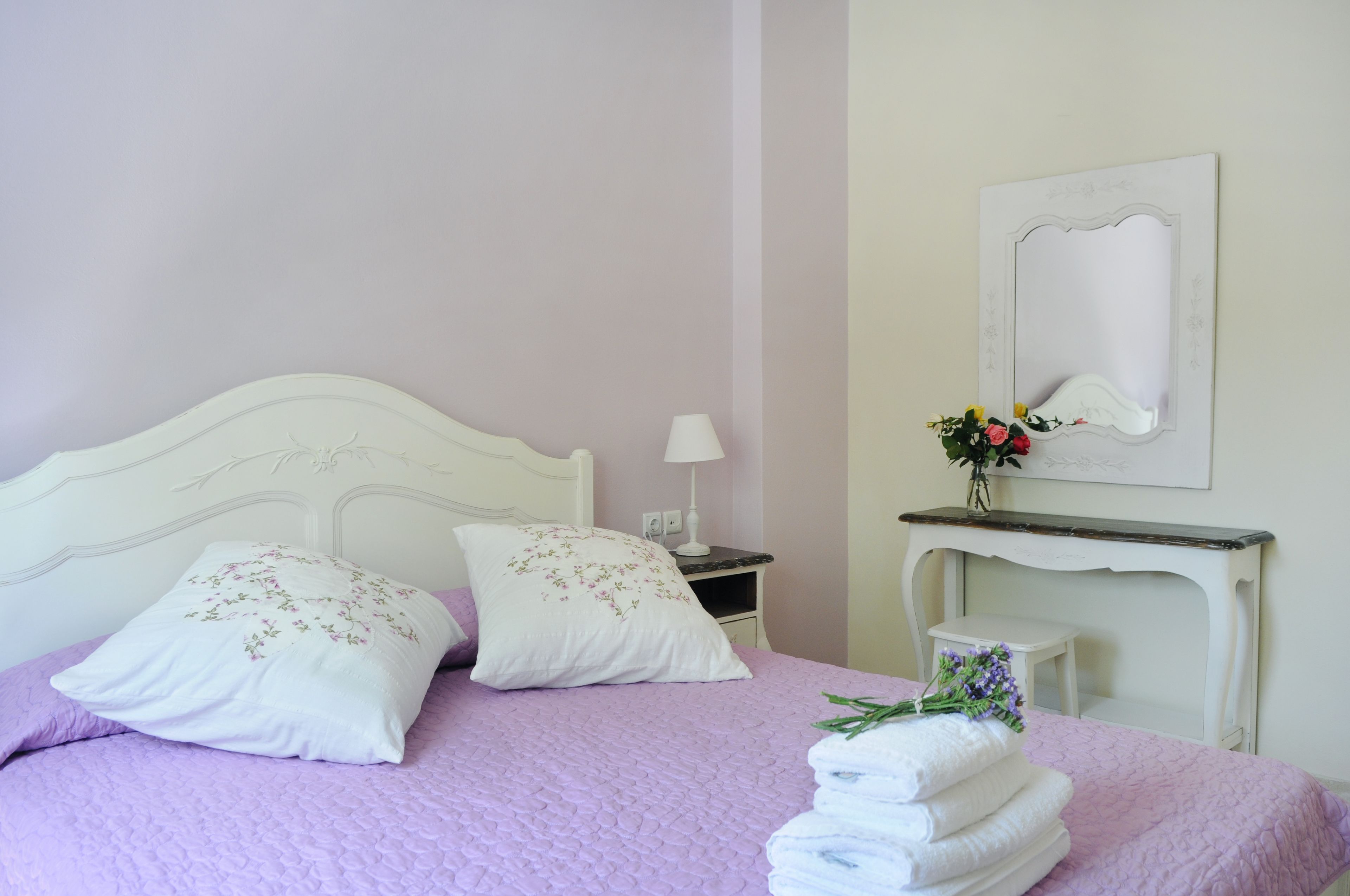 Een netjes opgemaakt bed met een lila deken, witte kussens met bloemenborduurwerk en een stap witte handdoeken.