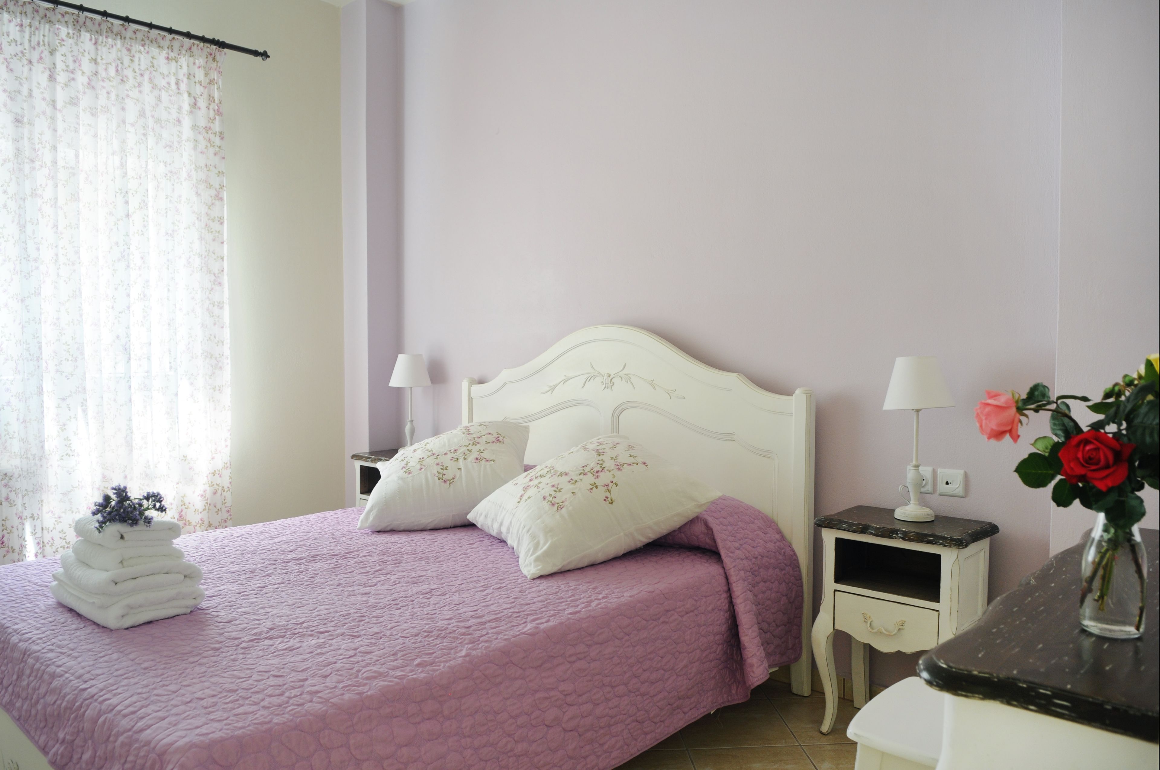 A neatly made bed with a pink bedspread and white pillows, a small bedside table with a lamp, and a vase with a red rose on the nightstand.