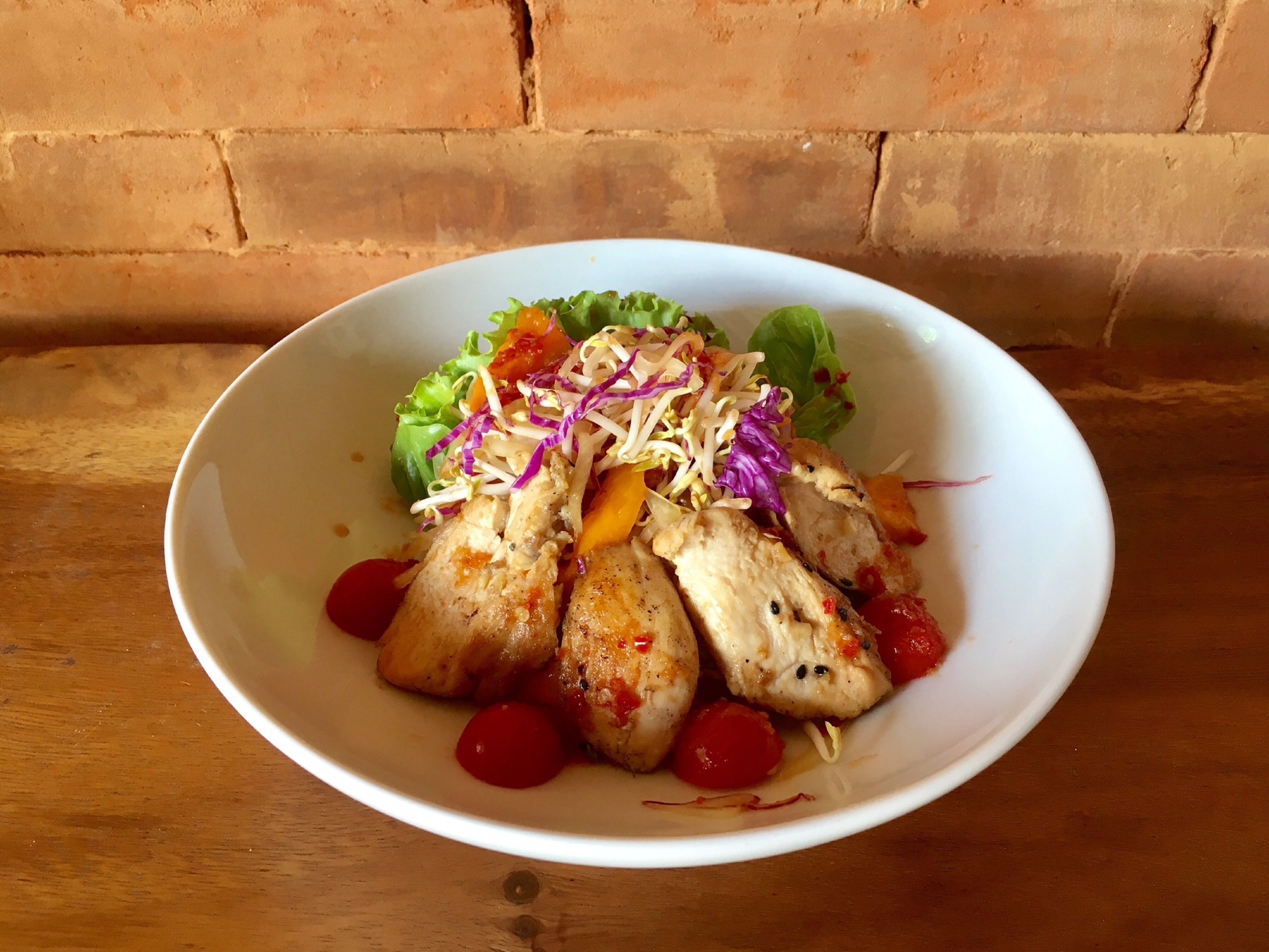 A white bowl with a salad containing lettuce, shredded cabbage, and cherry tomatoes, placed on a wooden table against a brick wall.