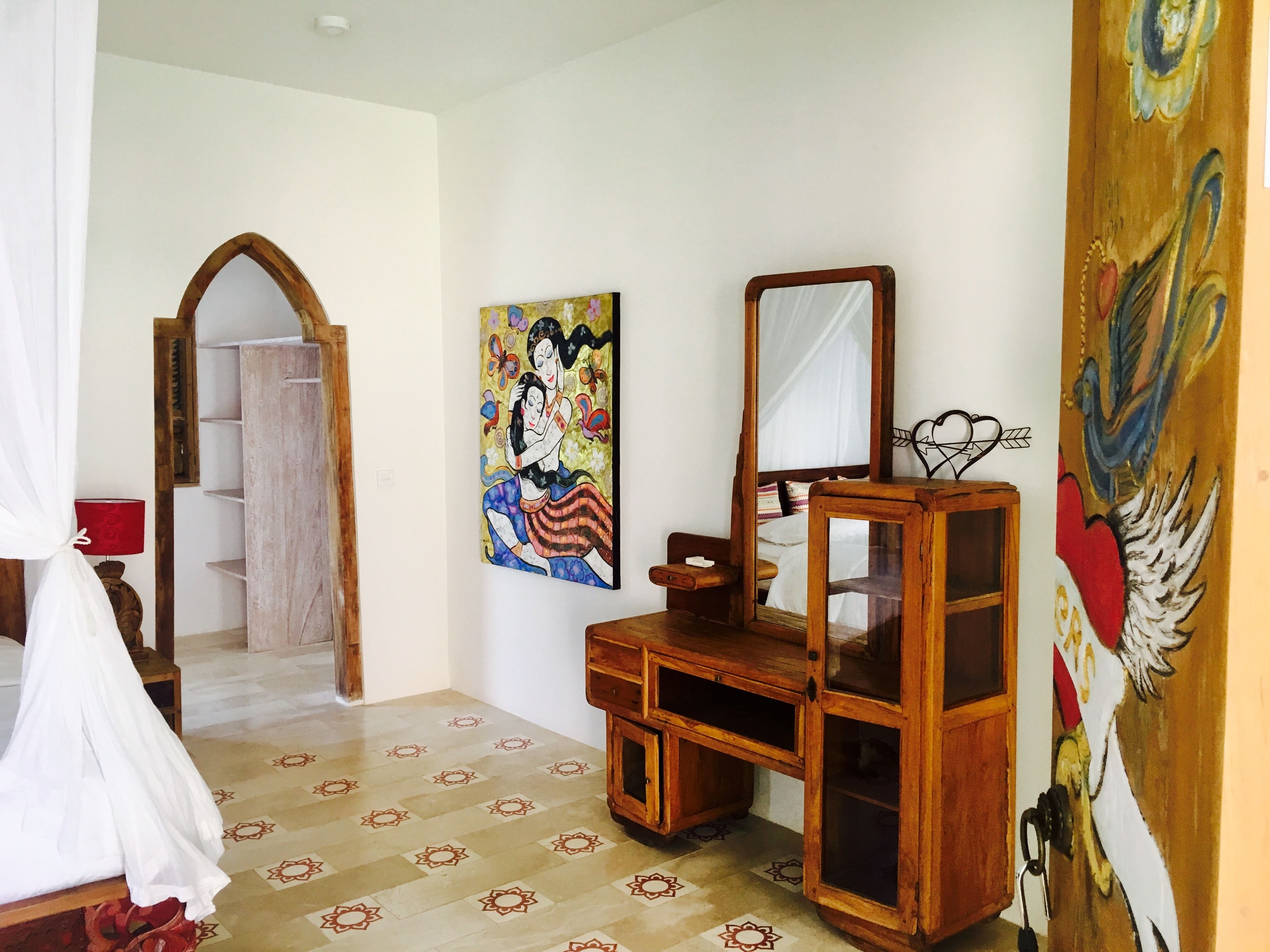 A bedroom with a bed, a wooden dresser, a mirror, a painting, and a door leading to another room.