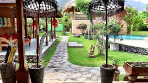 Un resort tropical con un sendero de piedra que lleva a una zona de piscina, rodeado de exuberante vegetación y construcciones de techo de paja.