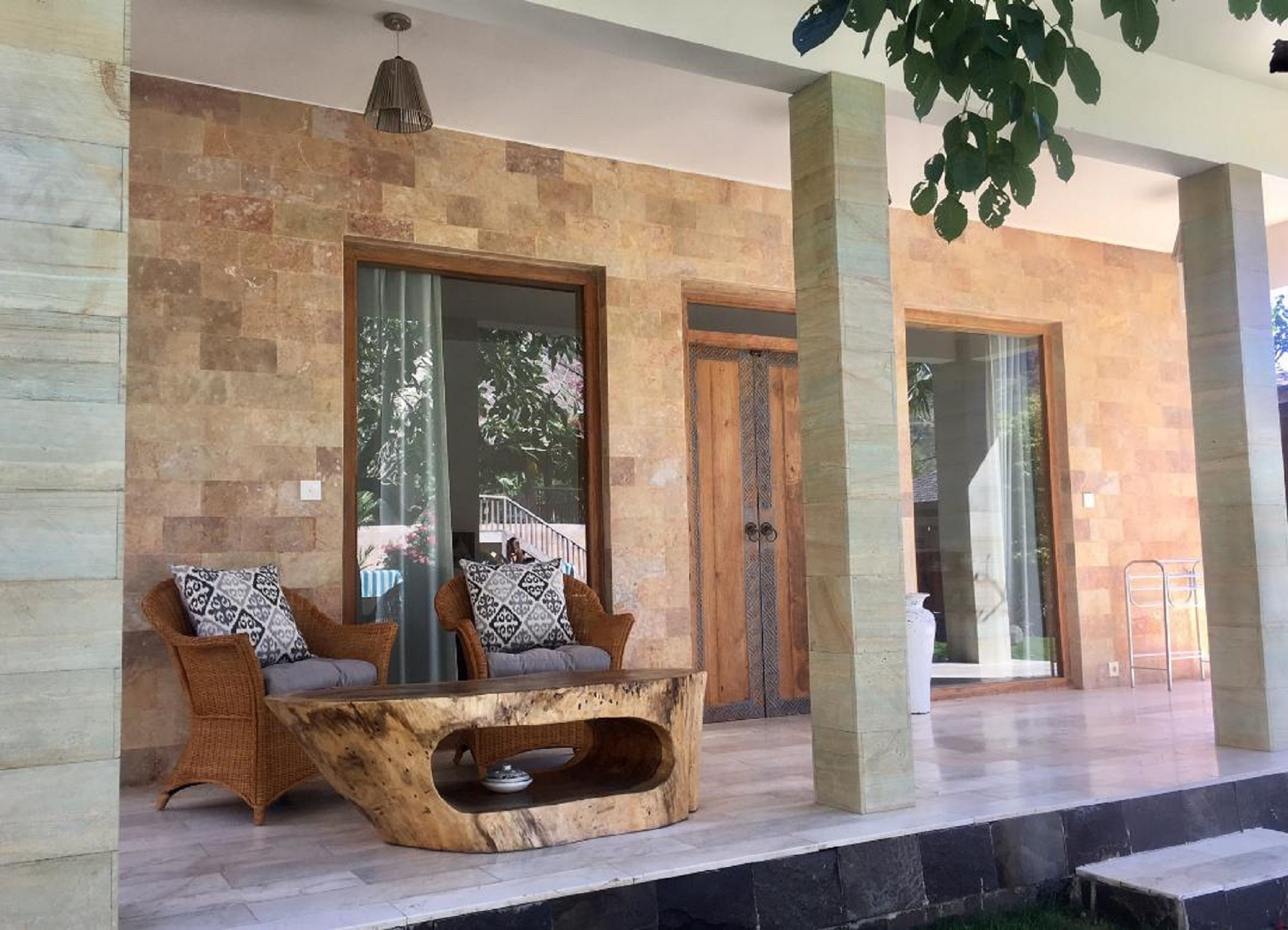 Un área de estar moderna al aire libre con mobiliario de mimbre y una mesa de madera original, frente a un muro de piedra con amplios ventanales que dan a un jardín.