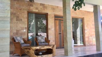 Un área de estar moderna al aire libre con mobiliario de mimbre y una mesa de madera original, frente a un muro de piedra con amplios ventanales que dan a un jardín.